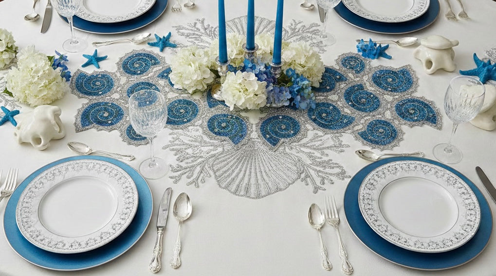 Elegant table setting with blue and white decor, including plates, cutlery, and floral arrangements on a white embroidered tablecloth with Handmade beaded decorative table runner in the shape of a shell with blue spiral design and silver rhinestone embellishments with silver carved cutlery and wine glasses and blue taper candles