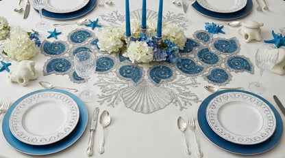 Elegant table setting with blue and white decor, including plates, cutlery, and floral arrangements on a white embroidered tablecloth with Handmade beaded decorative table runner in the shape of a shell with blue spiral design and silver rhinestone embellishments with silver carved cutlery and wine glasses and blue taper candles