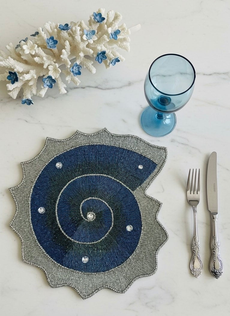 Handmade beaded decorative placemat in the shape of a shell with blue spiral design and silver rhinestone embellishments with silver carved cutlery and blue wine glass and white coral on a marble table
